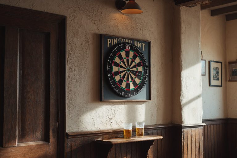 Dartbord in een donkere houten lijst aan de gepleisterde muur van een gezellige pub, met twee glazen bier op de plank eronder